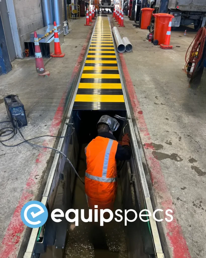 EquipSpecs technician welding inside a heavy vehicle inspection pit during the installation of automatic pit covers at a New Zealand workshop, with yellow and black walk-on-rated pit cover panels visible above.